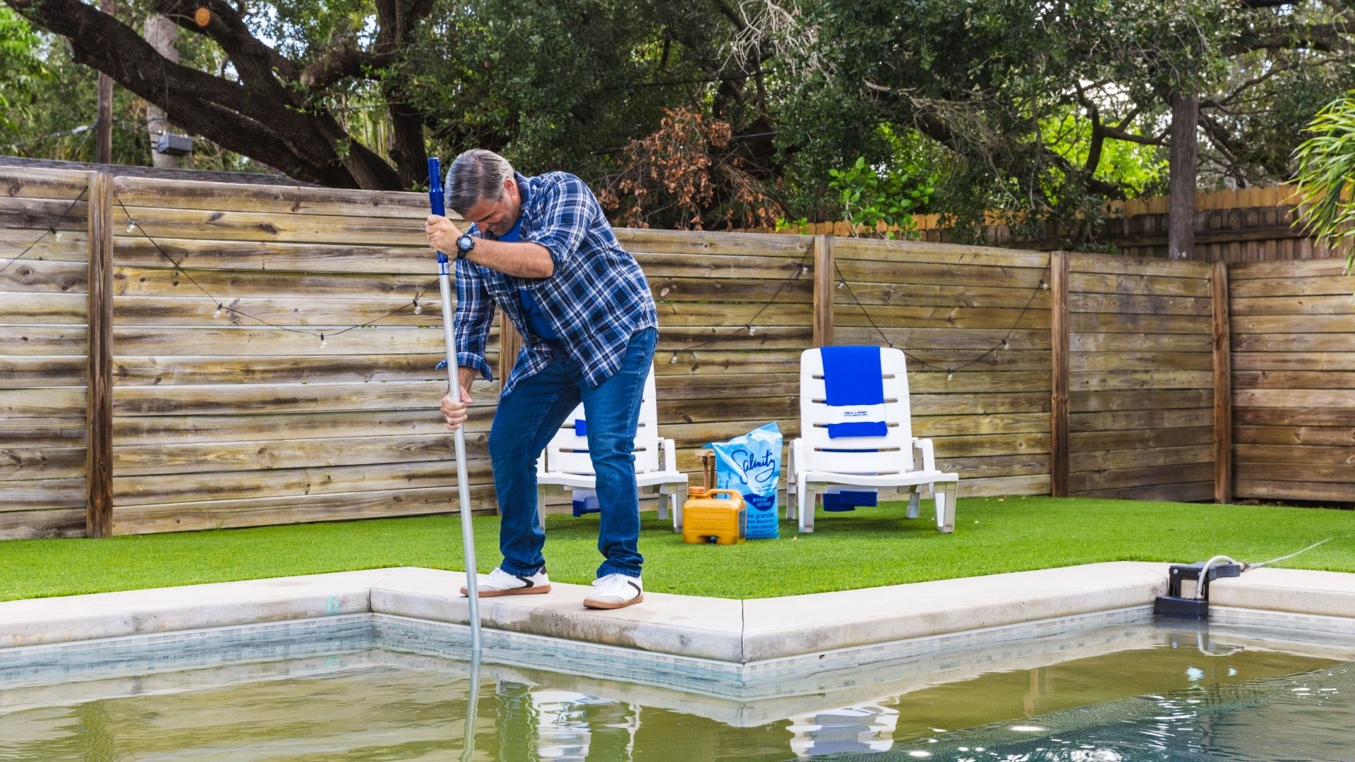 Middle-aged man in a blue plaid shirt brushing green, pollen-filled pool water with a telescopic pool brush along the pool edge. Two white lounge chairs with blue towels, a bag of pool salt, and a yellow chemical container sit on artificial turf behind him, bordered by a wooden privacy fence and trees.
