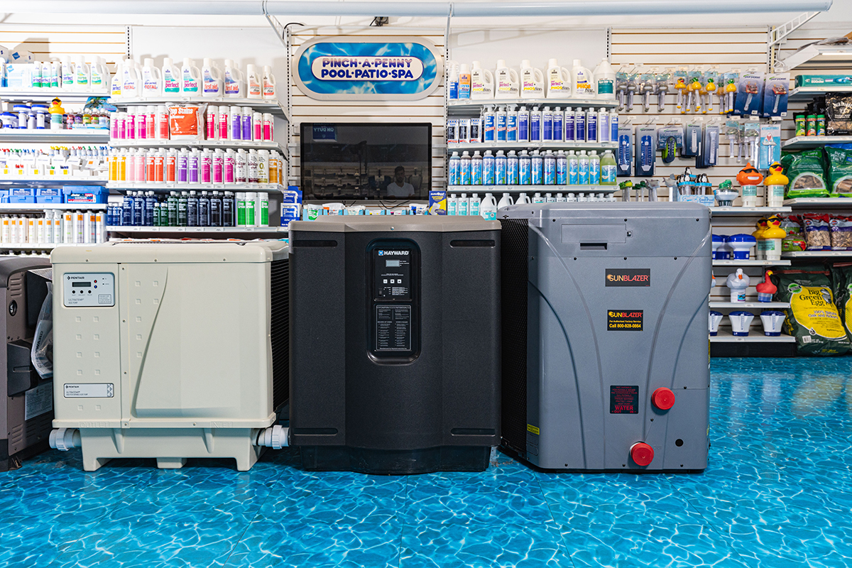 Three pool heaters displayed inside a pool supply store, including electric heat pump and gas-style units, positioned in front of shelves stocked with pool chemicals, testing supplies, and accessories.