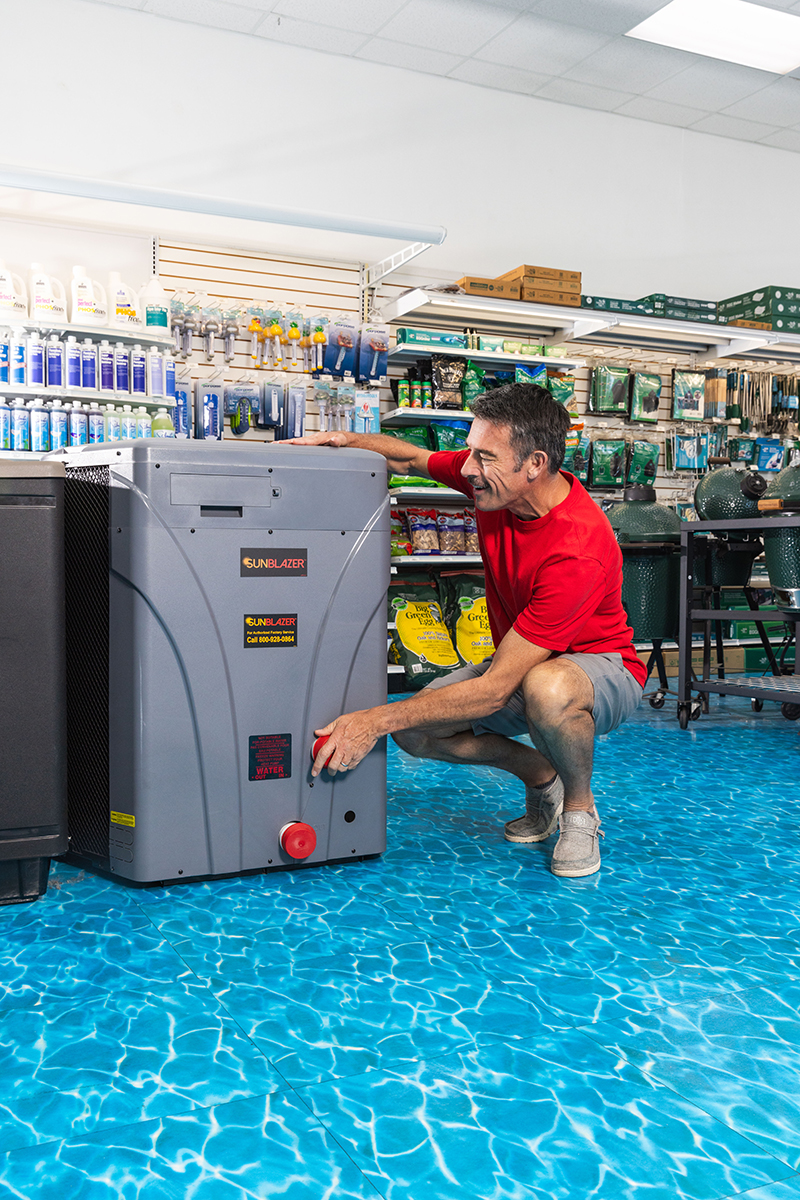 Customer kneeling inside a pool supply store, examining a SunBlazer pool heater and pointing to the water connection while surrounded by pool chemicals, equipment, and accessories on display.