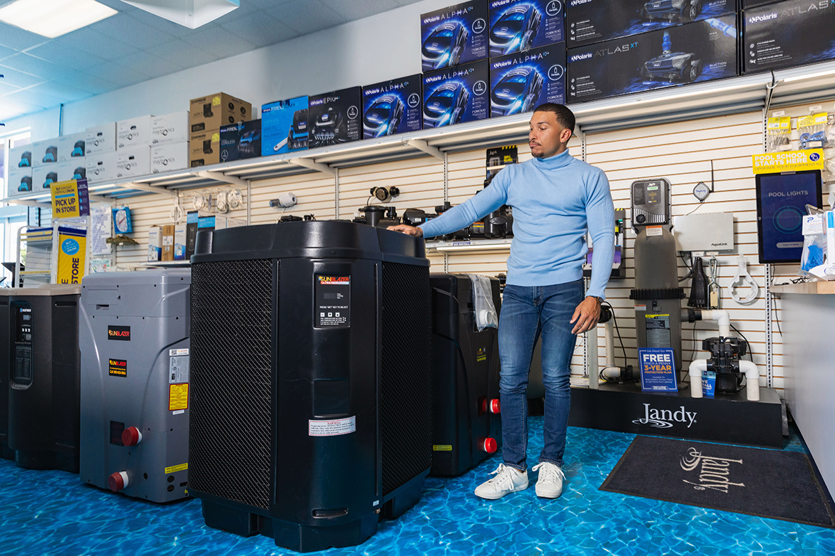 Customer standing inside a pool supply store, looking at a large SunBlazer pool heat pump alongside other pool heaters, pumps, and pool equipment on display.