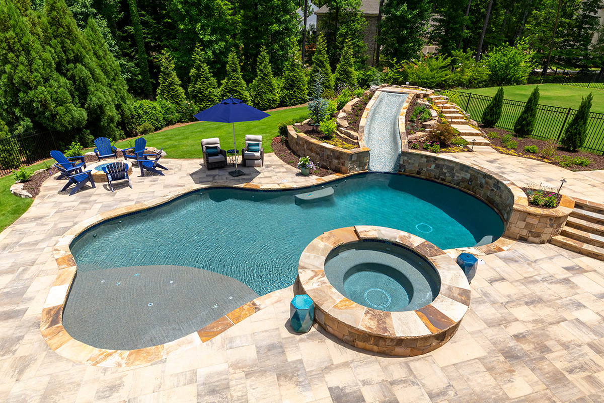 Freeform backyard swimming pool with an attached circular spa, stone coping, shallow sun shelf, and a raised water feature, surrounded by paver decking, lounge seating with umbrella, and lush landscaped greenery.