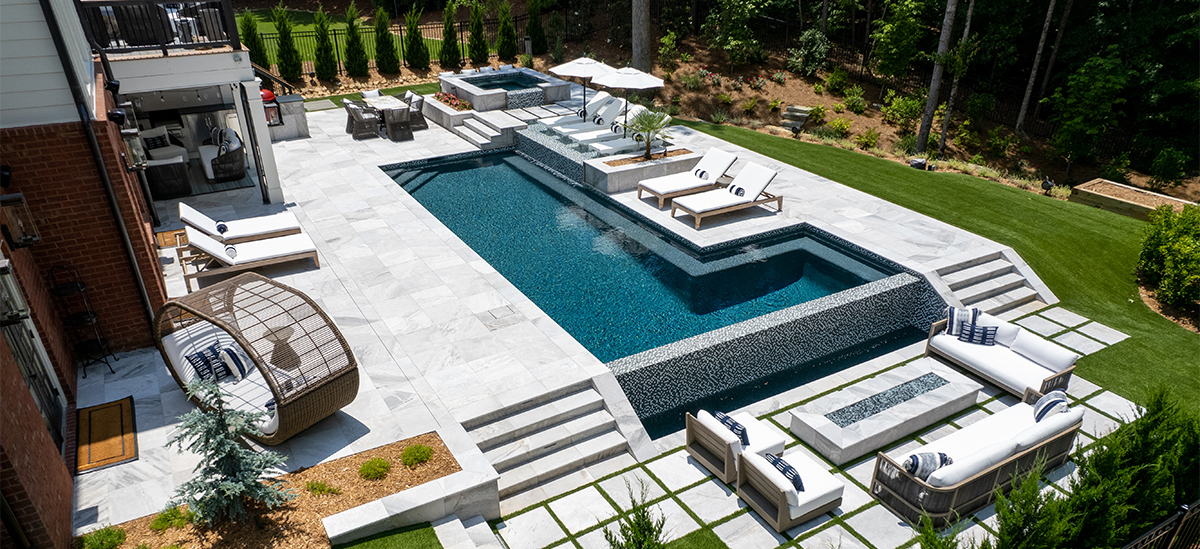 Modern backyard pool design featuring a long rectangular swimming pool with integrated spa, light stone decking, tiered steps, and multiple seating areas with lounge chairs, umbrellas, and outdoor furniture, set beside a multi-level home and landscaped lawn.